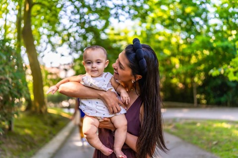 Happy latin mother smiling while holding her baby girl in a park, enjoying a sunny summer day