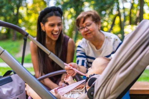 Grandmother feeds baby girl sitting in stroller, while mother watches, enjoying family time