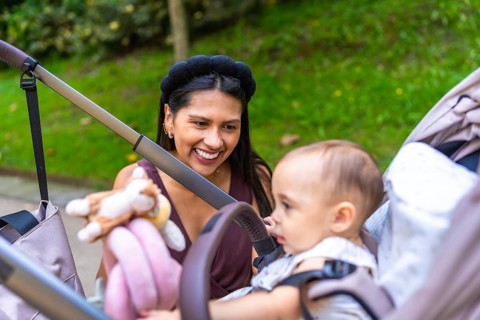 Happy latin mother entertaining her baby sitting in stroller at a park, enjoying family time