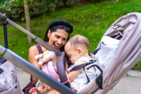Happy latin mother showing toys to her baby daughter sitting in a stroller, enjoying a sunny day