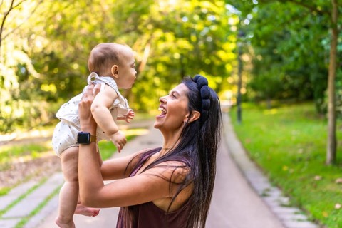 Smiling latin mother lifting her baby daughter while playing together in a sunny park, enjoying a