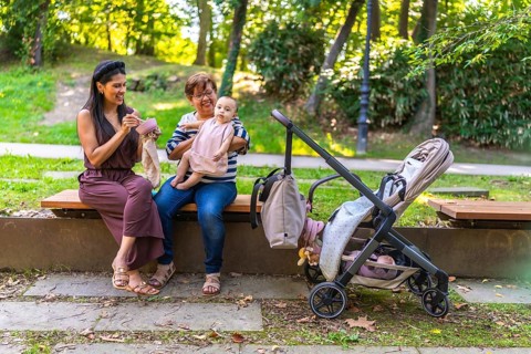 Grandmother holding baby girl while mother is eating and stroller is parked next to them on bench
