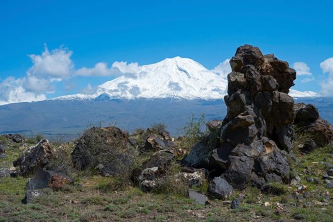 Snow-covered mountain under blue sky with clouds, surrounded by rocks and green landscape, aerial