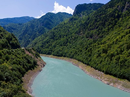 A turquoise-coloured river winds through a wooded and mountainous valley under a clear sky, aerial