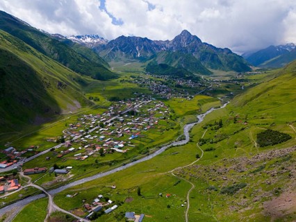Wide valley with river and village, surrounded by mountains under a partly cloudy sky, aerial view,