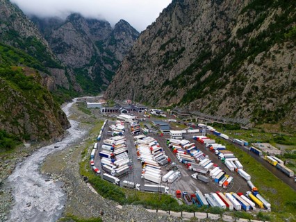 Car park full of lorries in a narrow mountain valley next to a river under a cloudy sky, aerial