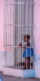 Little coloured girl behind window bars in a pink house, street of Trinidad, Cuba, Sancti Spíritus