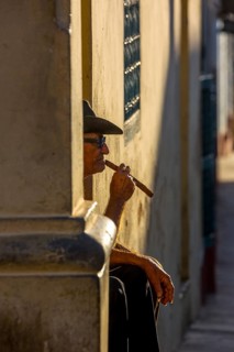 An old Cuban smokes a cigar and sits on a staircase in the warm light of the sunset, Trinidad,