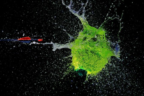 A green water balloon bursts powerfully under the impact of an arrow, against a dark background,