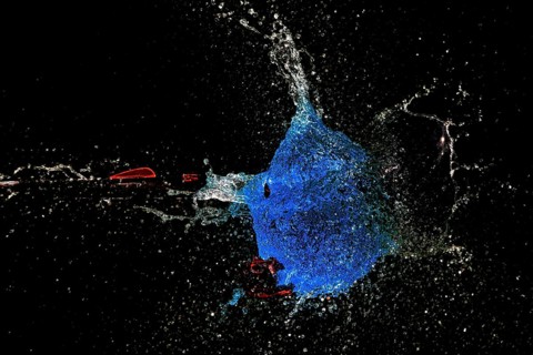 A blue water balloon bursts with force while an arrow shoots through it, surrounded by a dark