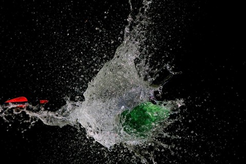A green water balloon bursts after arrow penetration, causing water to splash, in front of a dark