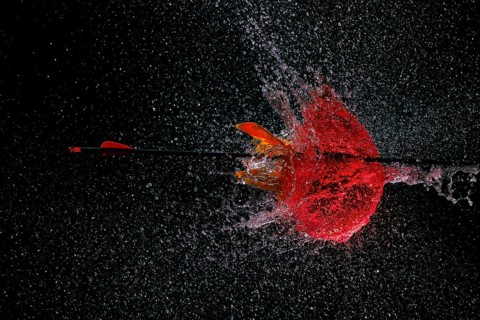 An arrow pierces a red water balloon and creates an impressive water explosion against a dark