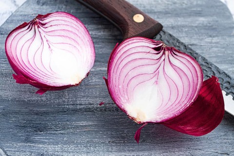 A vibrant red onion is cut in half, on a rustic wooden chopping board beside a sharp knife, ready