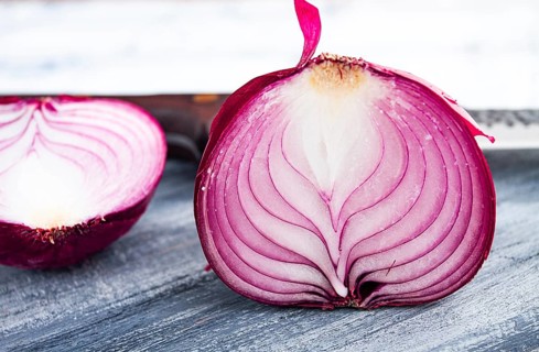 A vibrant red onion is cut in half, on a rustic wooden chopping board beside a sharp knife, ready