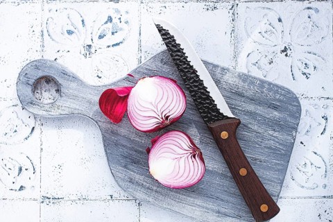 A vibrant red onion is cut in half, on a rustic wooden chopping board beside a sharp knife, ready