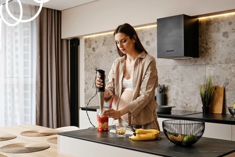 In a stylish kitchen, a pregnant woman blends fresh fruits like bananas and strawberries. Sunlight