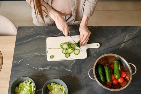 A pregnant woman with long hair skillfully slices cucumbers on a cutting board in a stylish kitchen
