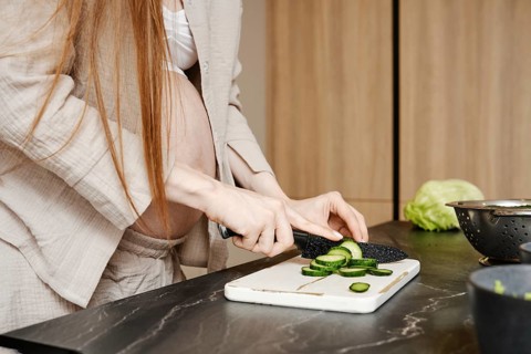 In a stylish kitchen, a pregnant woman with long hair cuts cucumbers on a white cutting board. She
