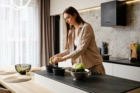 A woman with long hair is slicing vegetables in a contemporary kitchen. She focuses on her task