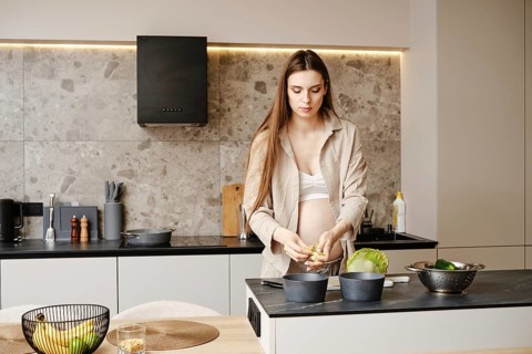 A pregnant woman stands in a stylish kitchen, chopping vegetables. She wears a casual outfit and
