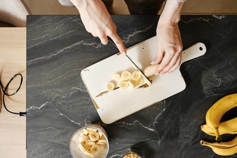 A person slices fresh bananas on a white cutting board in a well-lit kitchen. The countertop
