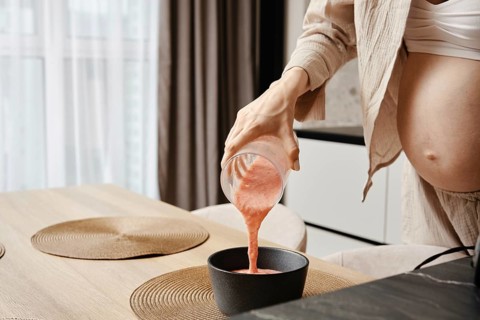 A pregnant woman pours a vibrant pink smoothie from a glass jar into a black bowl in a contemporary