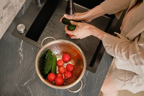 Hands scrub cucumbers under running water at a sleek kitchen sink. A colander nearby holds ripe