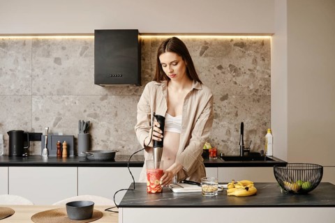 A pregnant woman stands in a contemporary kitchen using a handheld blender to prepare a fruit