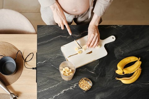 A person with a visible baby bump is cutting bananas on a wooden cutting board in a modern kitchen.