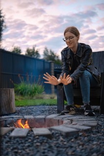 A young woman sits on an outdoor chair near a fire pit, warming her hands in the glow of the flames