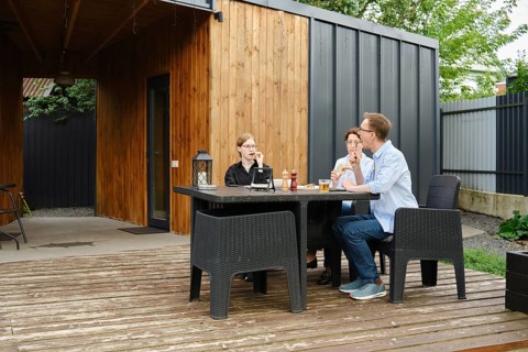 A family gather around a black table on a wooden deck of tiny house, sharing drinks and laughter in