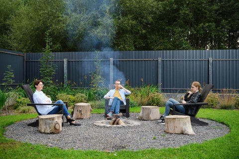 A family relax around a circular fire pit, seated on rustic chairs during a weekend. Smoke drifts