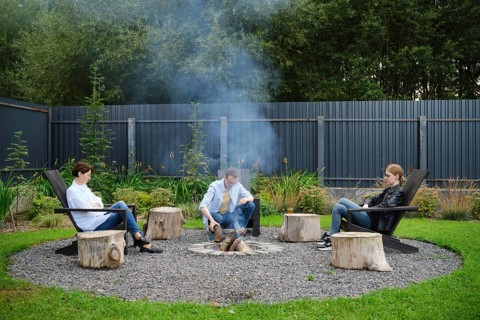A family sits comfortably on wooden chairs, gathering around a fire pit in a spacious garden. Smoke