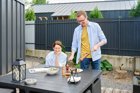 Middle-aged couple enjoy a barbecue meal outdoors in a backyard. Man is serving skewers on a grill,