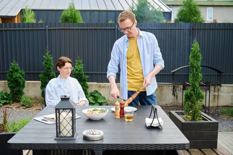 A man is serving grilled meat to a woman seated at a dining table in a beautiful backyard. The