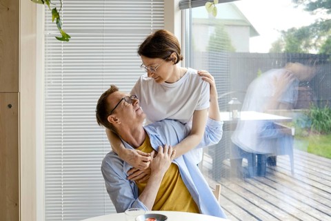 A man and woman share an affectionate moment indoors, surrounded by natural light. They smile at
