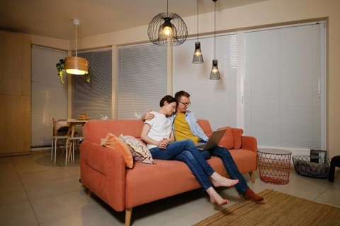 A husband embraces his wife while sitting on a couch in a modern living room. They are watching a