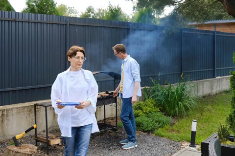 Two people are preparing a barbecue outdoors. A woman holds a plate while walking away, and a man