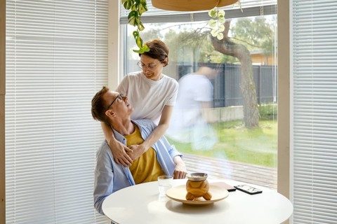 A couple shares a joyful interaction at a modern kitchen table. The woman embraces the man from