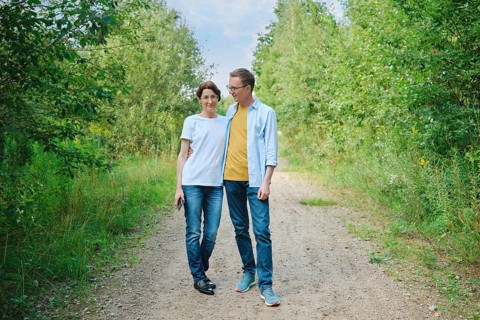 A middle-aged couple enjoys a stroll along a gravel path shaded by lush greenery. They smile at