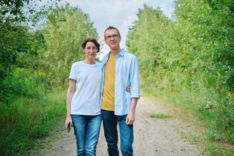 A middle-aged couple stands closely together on a natural path surrounded by lush greenery. They