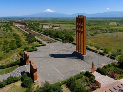 Large modern monument in front of a mountain panorama under a clear blue sky, aerial view,