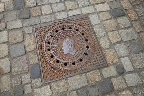 Square manhole cover with city coat of arms, inscription, paving stones, floor, African head with