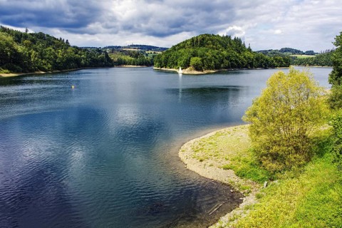 A lake (Aggertalsperre) with a wooded island and trees on the shore under a summer sky,