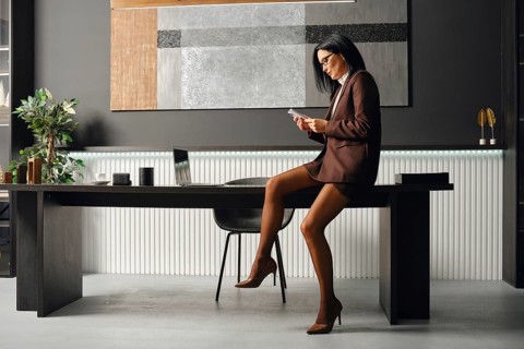 Serious business woman in a brown suit sits on a desk, focused on her smartphone. She checks her