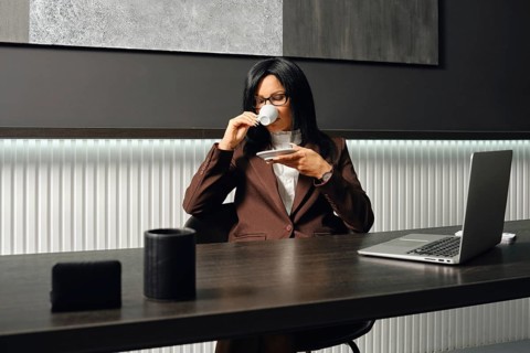 A real estate agent in a brown suit sits at a desk, sipping coffee from a small cup in a