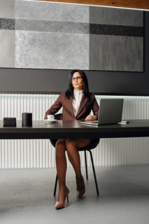 A slender woman with glasses and a stylish suit sits at an office desk with opened laptop. She