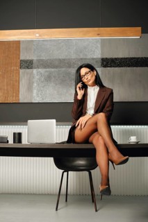 A confident businesswoman with slender legs in high hells sits on a tabletop of an office desk,