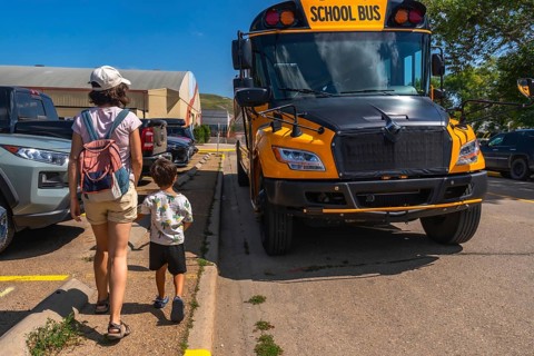 Mother and son walking hand in hand towards a bright yellow school bus in a bustling parking lot,