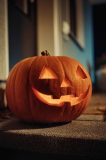 A carved, glowing pumpkin with a smiling face stands on a wall in front of a house at night,